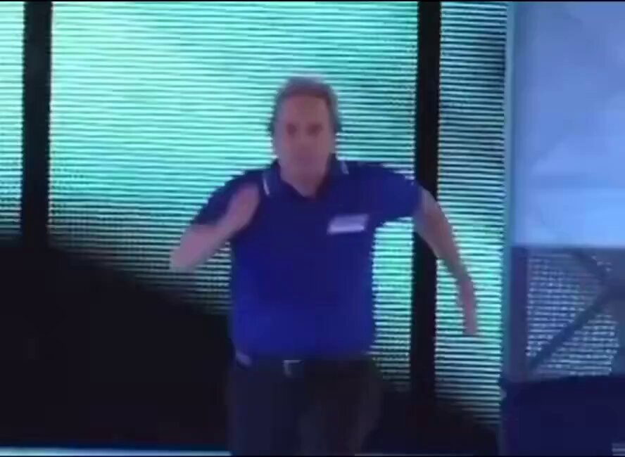 The image shows a man wearing a blue shirt, running across a stage with a large screen behind him displaying a message about Bitcoin. The background features a window that allows natural light into the room. The text "When you sign up on Club Orange and find out there are other Bitcoiners nearby." is visible in the image, suggesting that the man might be participating in an event related to Bitcoin or cryptocurrency.