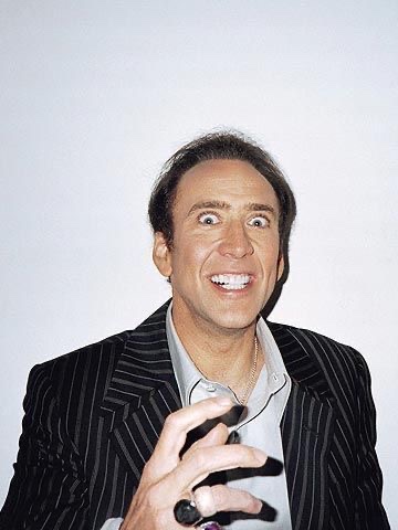 A man with receding brown hair, wearing a pinstripe suit jacket over a light shirt, makes an exaggerated wide-eyed expression while reaching his hand forward.