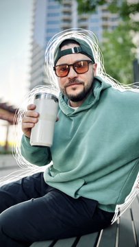 A man in a green hoodie, backwards cap, and sunglasses sits outdoors holding a reusable coffee cup, outlined with a white sketch effect.