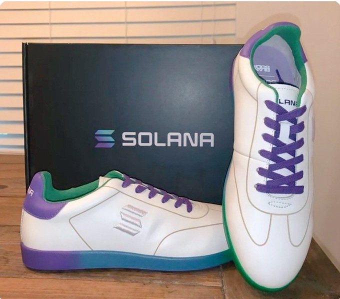 A pair of white sneakers with purple and green accents displayed next to a black Solana-branded shoebox.