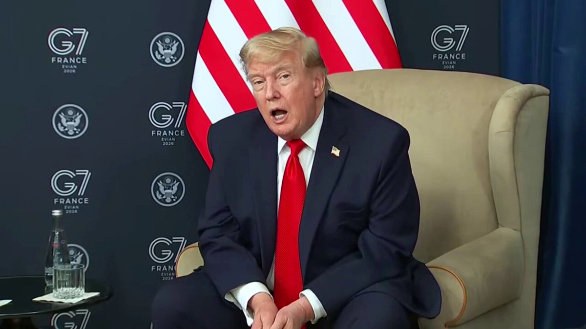 The image shows a man sitting in an armchair, wearing a suit and tie with his hands clasped together. He appears to be speaking or giving a speech, possibly addressing an audience at a political event. The setting includes a large flag behind him, which is part of a backdrop for the scene. There are also several other people present in the image, but they are not the main focus and do not appear to be directly involved with the man sitting in the chair.