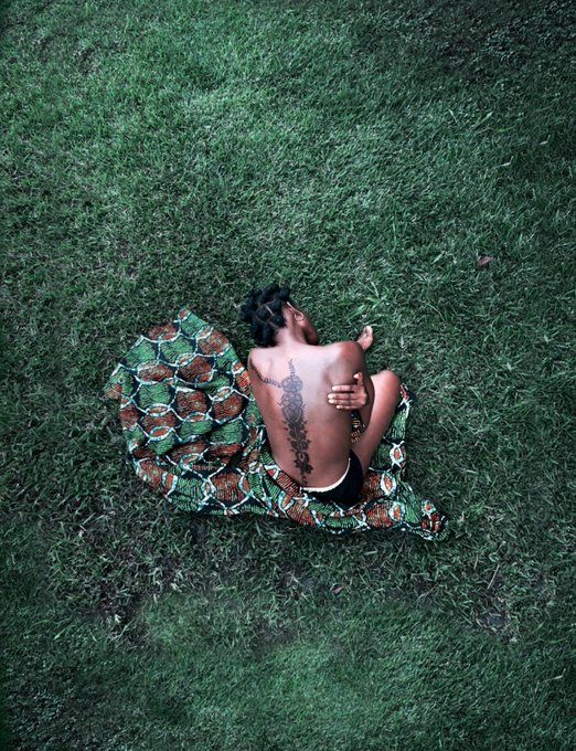 An overhead photograph of a shirtless person with dreadlocks and a back tattoo sitting on green patterned fabric spread over grass, embracing themselves.
