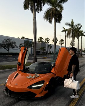 A woman poses next to an orange McLaren sports car (no visible text), with dihedral doors open, fitting the post's theme of newfound wealth from profiting on the $ANSEM meme coin.