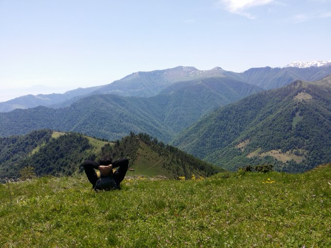 A scenic landscape photo of a person sitting in a grassy meadow with hands behind their head, overlooking lush green mountain ranges under a hazy sky.