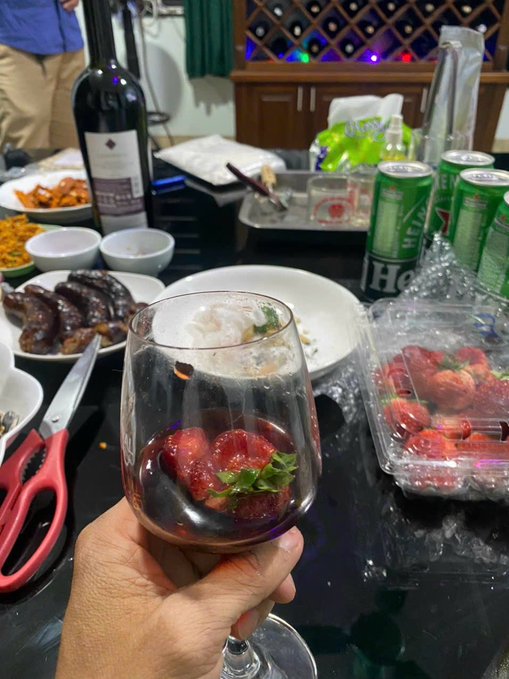 A hand holds a wine glass filled with fresh strawberries at a table spread with various dishes, a bottle of wine, Heineken cans, and more strawberries in a plastic container.