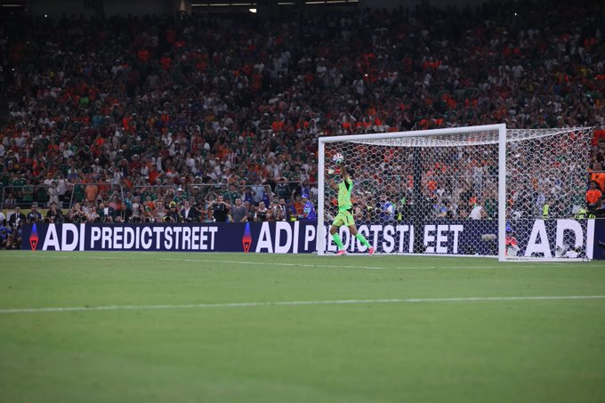 A soccer/football match photo showing a goalkeeper in green diving to save a ball, with a packed stadium crowd in the background and 'ADI PREDICTSTREET' advertising banners along the sideline.