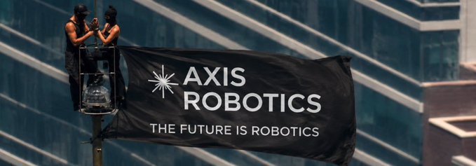 Two people on a platform attach a black flag reading "Axis Robotics - The Future is Robotics" to a pole in front of a skyscraper.