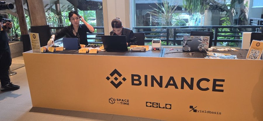 Two women staff a Binance-branded registration and merchandise booth at Binance Campus Asia 2026, with the event's front desk also featuring sponsor logos for SPACE and TIME, CELO, and yieldbasis alongside laptop check-ins and a QR code display for attendees.