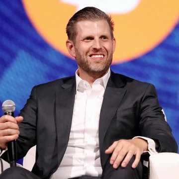 Eric Trump smiles broadly while seated on stage at what appears to be a panel or conference event, holding a microphone, illustrating his public denial of rumors about the Trump family launching a new meme coin.