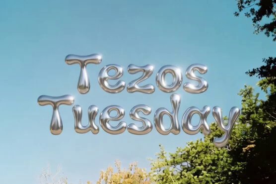 The image features a large, metallic balloon with the words "Tezos Tuesday" written on it in silver letters floating in the air against a clear blue sky backdrop. The balloon appears to be floating above some trees, adding an artistic touch to the scene. The text is positioned centrally and stands out prominently due to its size and color.