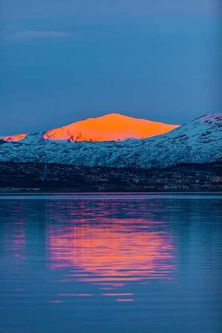 A snow-covered mountain peak glows vibrant orange at sunset, its reflection stretching across the calm blue waters below.