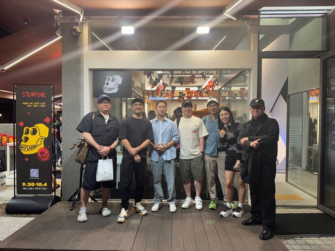 A group of seven people poses for a photo outside a brightly lit storefront featuring a 'STOMP' banner with a skull design, the date '9.30-10.4', and festive decorations.