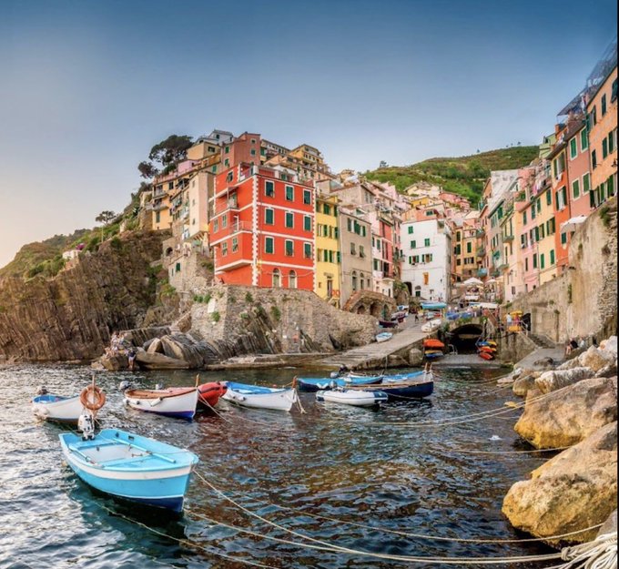 A picturesque coastal village with colorful houses (red, yellow, pink) built along rocky cliffs above a small harbor with several small boats moored in calm water.