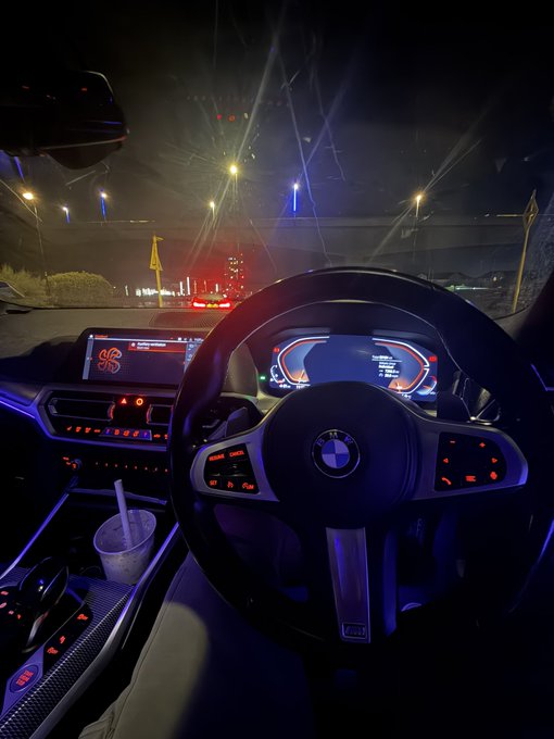 Nighttime view from the driver's seat of a BMW, with the dashboard and steering wheel illuminated in purple ambient lighting as the car is stopped on a road with streetlights and traffic ahead.