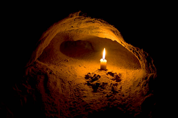 A single candle burns brightly inside a small cave-like rock alcove, casting a warm golden glow against the surrounding darkness, with shadowy paw prints visible in the sand below. The image metaphorically represents the post's message about "Lighter" ($LIT), where the solitary flame symbolizes an emerging opportunity that is still undiscovered by most investors who remain "underexposed" in the dark, waiting for them to eventually "buy higher."
