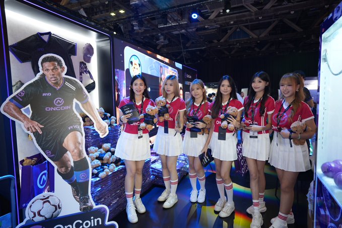 Six women in red-and-white promotional outfits stand beside a large OnCoinLive display featuring a footballer cutout, holding small plush toys at the TOKEN2049 event. Visible branding includes “OnCoin,” “OnCoin Live,” “RIO,” and “UNCOM,” matching the post’s description of visiting the OnCoinLive booth for a live stream.