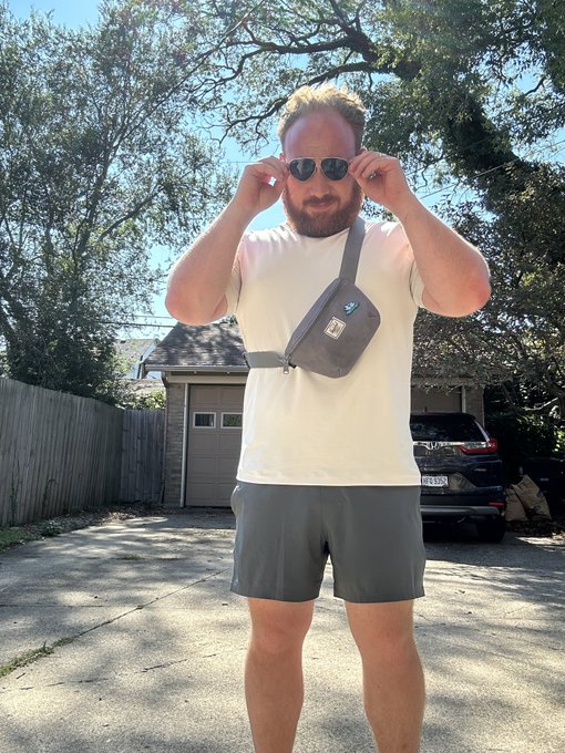 A bearded man standing in a driveway wearing a cream t-shirt, gray shorts, a BMW crossbody bag, and adjusting his sunglasses, with a house and car visible in the background.