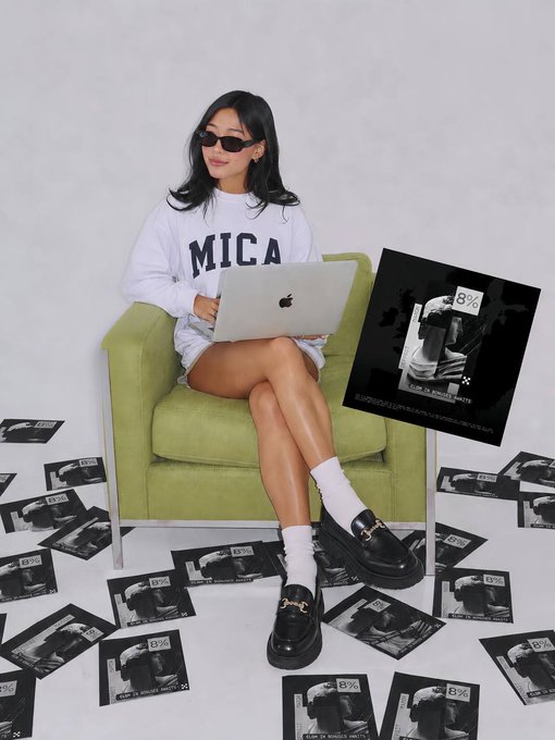 A young woman wearing a white 'MICA' sweatshirt and sunglasses sits on a green chair using a MacBook, surrounded by scattered copies of an '8%' magazine/zine featuring sneaker photography.