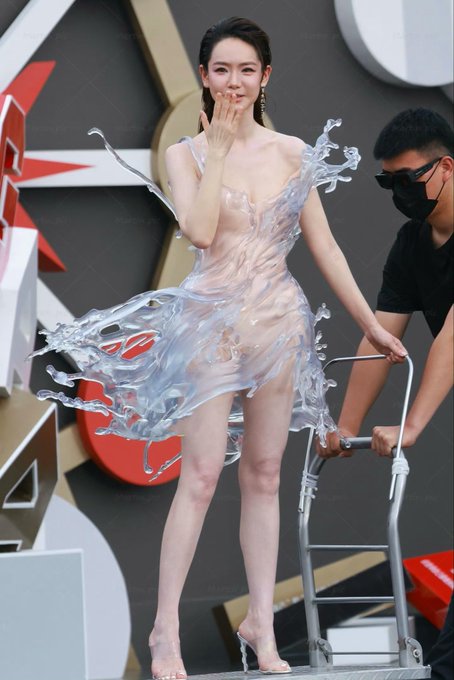 An Asian woman wearing an elaborate sheer, sculpted dress waves while descending a small ladder, assisted by a masked man.