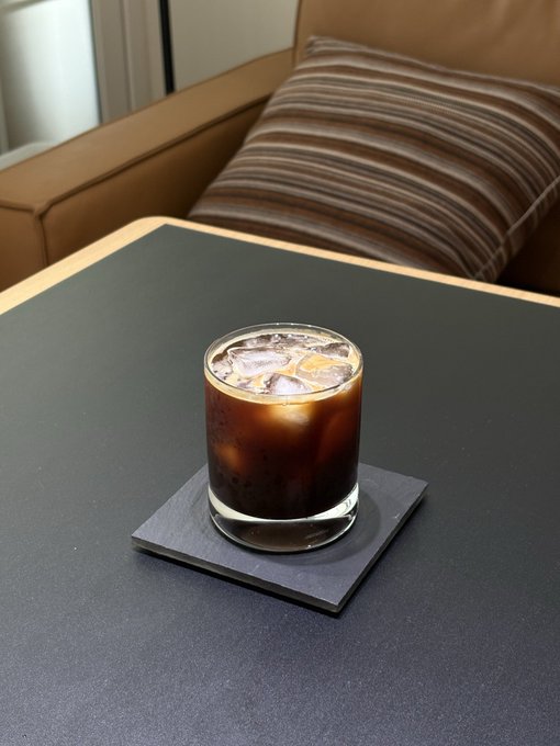 A glass of iced coffee with ice cubes sits on a black slate coaster atop a dark table, with a brown striped pillow visible in the background.