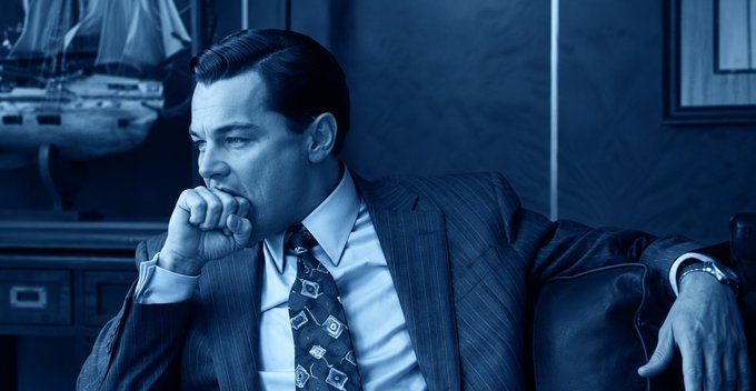 A blue-tinted photograph of a man in a suit and tie, sitting in a thoughtful pose with his hand over his mouth, with a model ship in the background.