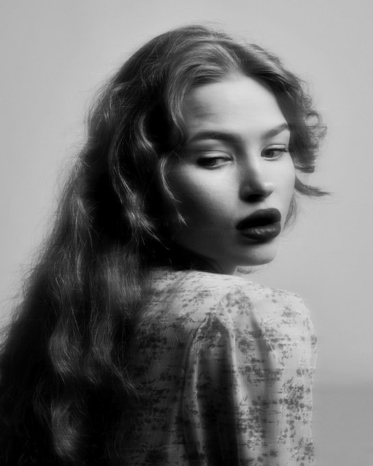 A black and white portrait photograph of a woman with long wavy hair looking over her shoulder, wearing a floral patterned top, against a plain background.