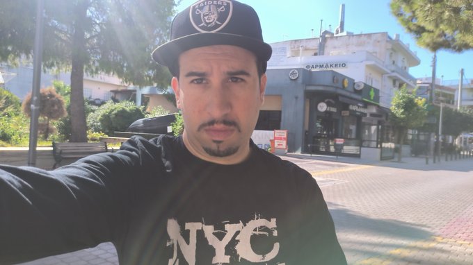 A man wearing a black Raiders cap and an "NYC" sweatshirt takes a selfie outdoors, with a "ΦΑΡΜΑΚΕΙΟ" (pharmacy) storefront visible in the background.