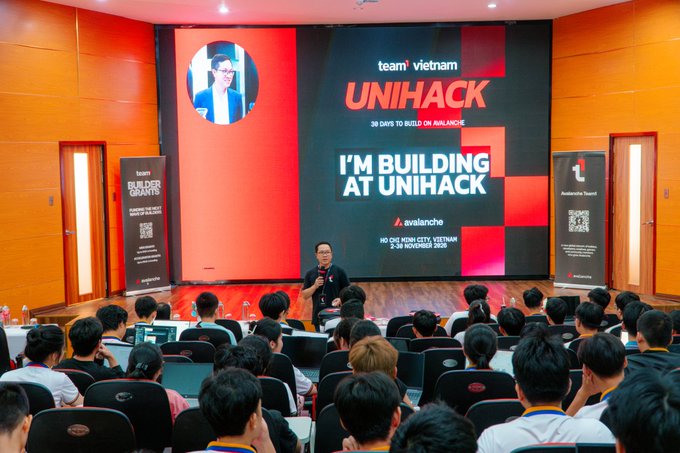 A conference setting where a presenter stands on stage in front of a large screen displaying 'team vietnam UNIHACK', 'I'M BUILDING AT UNIHACK', and 'Avalanche', with an audience seated in the foreground.
