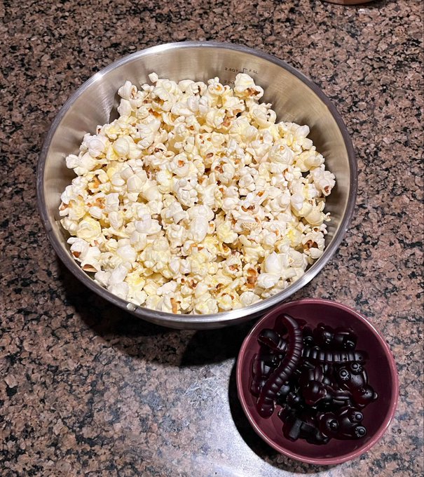 A metal bowl filled with freshly popped popcorn sitting on a granite countertop next to a small red bowl containing dark red gummy bears.