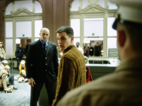 A man in a brown sweater looks back suspiciously at someone, while another man in a dark suit stands beside him, with people sitting on the floor in what appears to be a train station or terminal setting. The accompanying post suggests the image relates to claims of coordinated online misinformation campaigns, with the tense, watchful expressions of the characters in the image serving as a fitting visual metaphor for the idea of a coordinated group acting suspiciously.