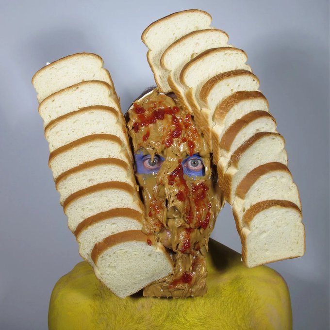 A surreal portrait of a person with a face covered in what appears to be peanut butter and jelly, framed by slices of bread arranged like wings, against a gray background.