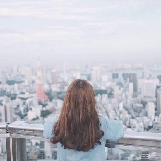 A woman with long brown hair wearing a light blue shirt looks out over a hazy city skyline featuring Tokyo Tower from an observation deck.