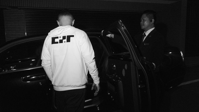 Black-and-white photo of a man in a white "HJC" hoodie getting into a car, assisted by a man in a suit holding the door open.