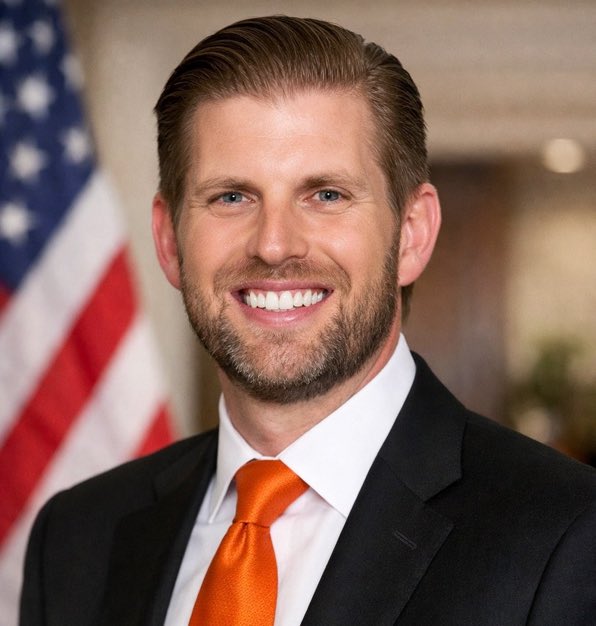 Professional headshot of a smiling man with a beard wearing a dark suit, white shirt, and orange tie, standing in front of an American flag.