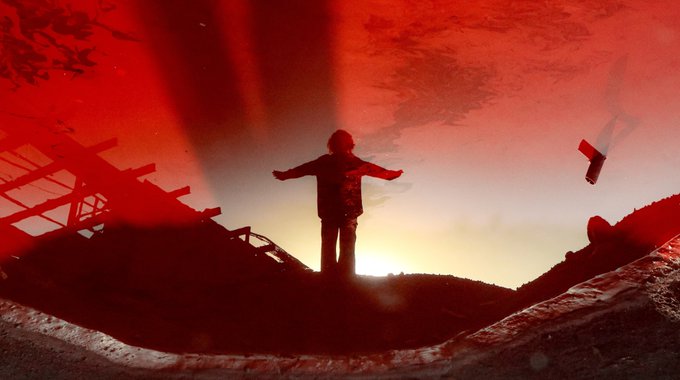 A silhouetted figure with outstretched arms stands against a dramatic red and orange sunset, with a small flag visible to the right.