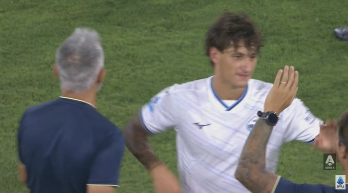 A screenshot from a Serie A soccer match showing a player in a white Lazio jersey with the Diadora logo, celebrating near a coach with gray hair on the sideline.
