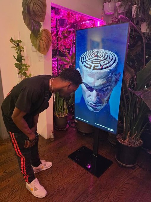 A young man with a curly fade haircut stands in a plant-filled room with pink grow lights, smiling as he leans toward a digital display showing artwork of a classical bust topped with a maze pattern.