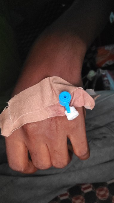 A person's hand with an IV cannula secured by a tan cloth bandage and a blue medical clip rests on gray fabric. The post suggests the writer is hospitalized or receiving medical treatment, advising followers to prioritize self-care like sleep, nutrition, hydration, and exercise.