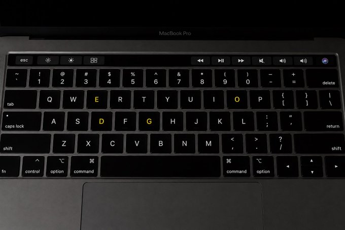 A MacBook Pro keyboard is shown with the letters "E," "D," "O," and "G" illuminated in yellow, spelling out "DOGE" across the keys. This visual cue connects to the cryptocurrency ticker $DOGE, using the keyboard's backlight feature to spell out the token's name.