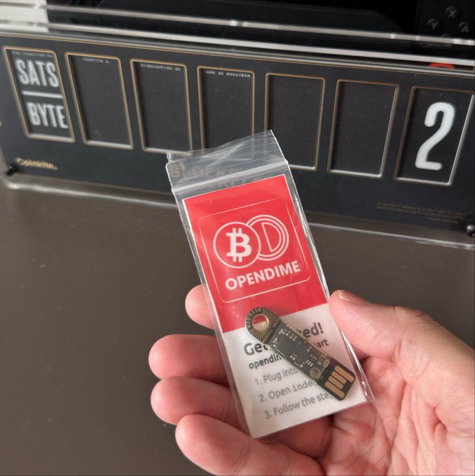 A hand holds an OpenDime Bitcoin hardware wallet in its original packaging against a vintage satellite scoreboard background displaying "SATS BYTE" and the number "2." The product tag invites users to "Plug in," "Open index," and "Follow the steps," prompting the joke about creative misuses for the secure crypto storage device.