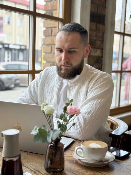 A bearded man in a white knit cardigan works on a laptop at a cafe, seated near a window with a cappuccino, flowers, and a phone on the wooden table.