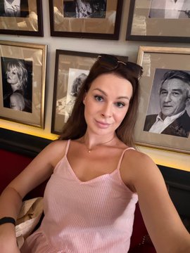 A selfie of a young woman with long brown hair wearing a pink tank top, seated in front of a wall decorated with framed black-and-white portraits.