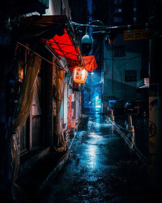 A moody, rain-soaked city alley at night illuminated by a glowing red paper lantern, with Korean signage including '다목적 CCTV' (CCTV) and a 'Z' logo visible on a building.