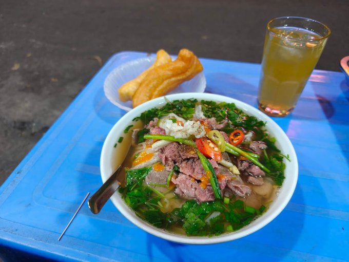 A bowl of Vietnamese pho with beef, herbs, and chili peppers sits on a blue plastic table alongside fried dough sticks and a glass of iced tea.