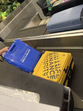 Two toy trucks labeled "BINANCE" sit on a baggage carousel at an airport, fitting the post's "taking off, next stop, turning things around" travel theme.