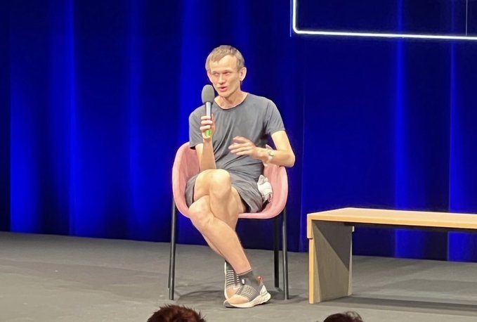 A man in a gray t-shirt and shorts sits in a pink chair on a stage with blue curtains, holding a microphone and speaking, with a wooden bench beside him.