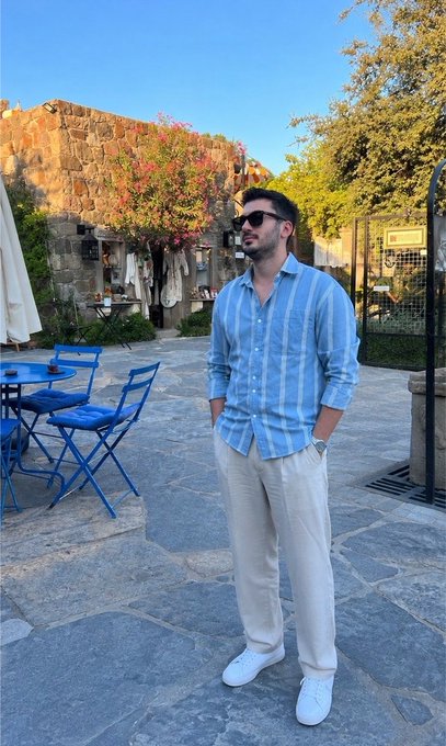 A man wearing sunglasses, a blue striped shirt, and white pants stands in a charming stone courtyard with blue café chairs and flowering vines, exuding a relaxed vibe that complements his cheerful "Good morning" greeting wishing followers a great day.