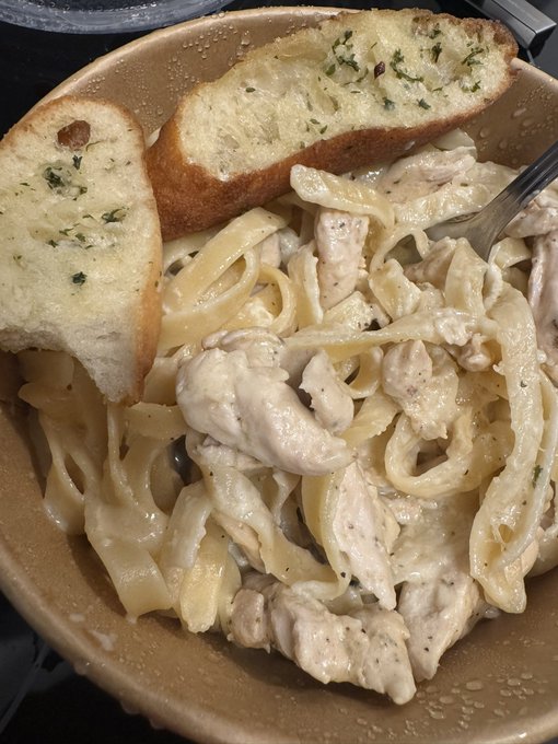 A bowl of creamy chicken alfredo fettuccine pasta served in a kraft takeout bowl with two slices of garlic bread on top, perfect for a satisfying comfort food craving. The pasta is generously coated in a rich white sauce with visible chunks of chicken, ready to be enjoyed straight from the container.