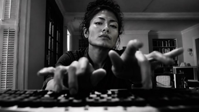 A dramatic black-and-white photo shows a woman with dark hair leaning over a chessboard with her hands outstretched toward the camera in an intense gesture.