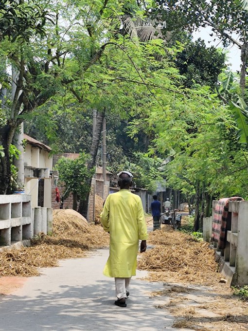 The image shows a man walking down an alleyway, wearing a yellow outfit that stands out against the surroundings. The alleyway appears to be filled with hay or straw, which could indicate that the photo was taken during a time of transition between seasons. The man is walking away from the camera and seems to be heading towards something in the distance.